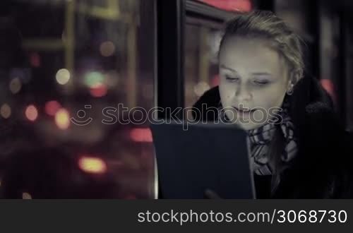 Young woman watching video on touchpad, then choosing another one during evening ride in the bus. Defocused city traffic in the bus window