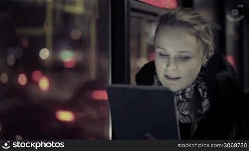 Young woman watching video on touchpad, then choosing another one during evening ride in the bus. Defocused city traffic in the bus window