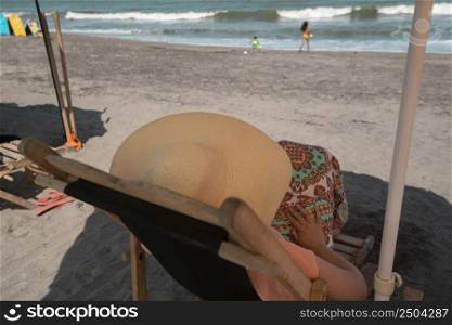 Young woman wearing a hat and dress resting on a wooden chair, keeping an eye on her children who are playing on the beach during a sunny morning. Young woman wearing hat and dress resting on a wooden chair on the beach with the sea in the background during a sunny morning