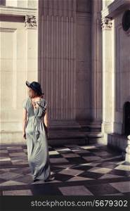 Young woman wearing an elegant dress and hat is standing in an impressive and majestic hall bathed in sunlight