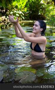 Young woman wearing bikini kneeling in pond, arms raised, cupping water in hands