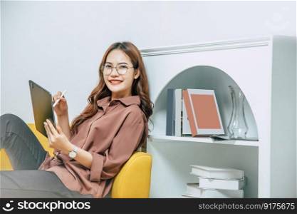 Young woman wearing glasses sitting on sofa and using technology of digital pen on tablet to working from home during self-isolation and quarantine covid-19 coronavirus outbreak