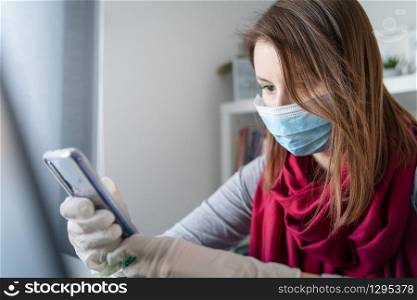 Young woman wearing protective gloves on hands and mask on face working from home or at office work by the table laptop mobile phone texting sms preventing virus spread epidemic quarantine prevention