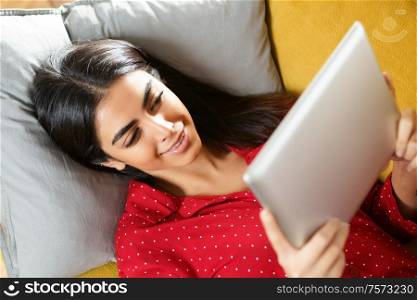 Young woman wearing pyjamas at home using digital tablet on a couch. Persian woman at home using digital tablet