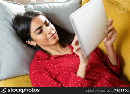 Young woman wearing pyjamas at home using digital tablet on a couch. Persian woman at home using digital tablet