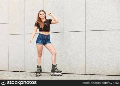 Young woman wearing roller skates standing against concrete wall in city. Female being sporty having fun during summer time.. Young woman riding roller skates