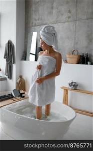 Young woman wearing white towel on head and body coming out of bath. Morning body care, skincare in bathroom concept. Young woman wearing white towel on head and body coming out of bath