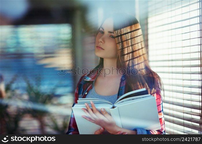 Young woman with a book standing near window  shot through window glass with reflections and flares