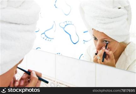 Young woman with a towel draped around her hair, putting on eye liner in front of the mirror