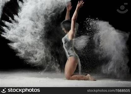 young woman with an athletic body sits on the floor and throws white flour up, dust flies in different directions, an athlete is dressed in a black bodysuit