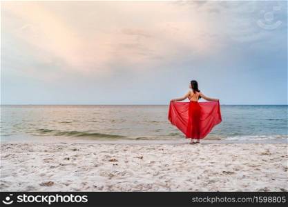 young woman with arms spread in a red dress on the sea beach with wind blow
