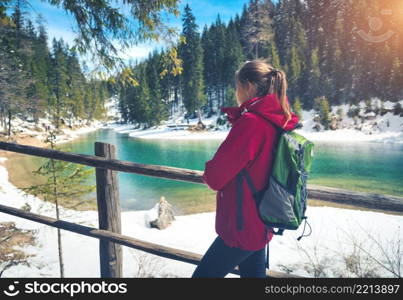 Young woman with backpack is standing near lake with blue water at sunny day in spring. Travel. Landscape with slim girl, wooden fence, reflection in water, snowy mountains, orange trees in winter