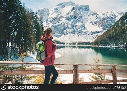 Young woman with backpack is standing near lake with blue water at sunny day in autumn. Travel. Landscape with slim girl, wooden fence, reflection in water, snowy mountains, orange trees in fall