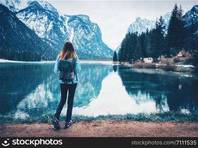 Young woman with backpack is standing on the coast of mountain lake at cloudy day in autumn. Travel in Italy in fall. Landscape with slim girl, reflection in water, snowy rocks, trees. Vintage toning