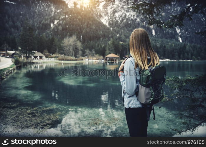 Young woman with backpack is standing on the coast of mountain lake at sunset in autumn. Travel in Italy. Landscape with slim girl, reflection in water, snowy rocks, green trees in fall. Vintage
