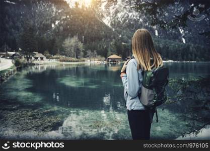 Young woman with backpack is standing on the coast of mountain lake at sunset in autumn. Travel in Italy. Landscape with slim girl, reflection in water, snowy rocks, green trees in fall. Vintage