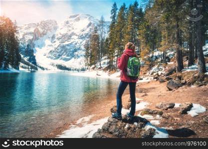 Young woman with backpack is standing on the stone near lake with azure water at sunny day in autumn. Travel. Landscape with slim girl, reflection in water, snowy mountains, orange trees in fall