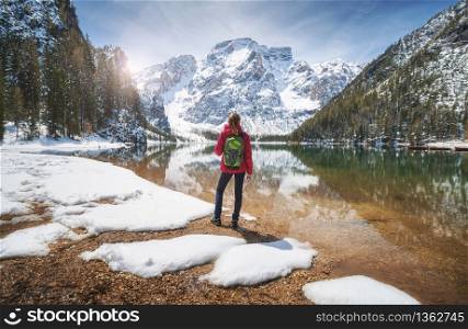 Young woman with backpack on the snowy shore of Braies lake with clear water at sunny bright day in spring. Travel. Landscape with slim girl, reflection in water, mountains, green trees, blue sky