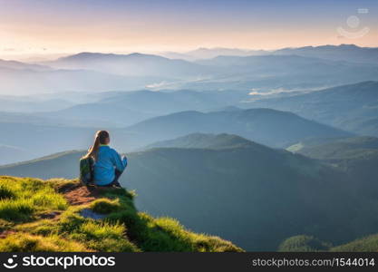 Young woman with backpack sitting on the mountain peak and beautiful mountains in fog at sunset in summer. Landscape with sporty girl, green grass, forest, hills , sky, sunbeams. Travel and tourism