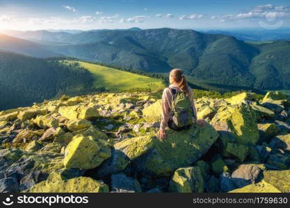 Young woman with backpack sitting on the stone on mountain peak. Beautiful mountains at sunset in summer. Landscape with relaxing sporty girl, green stones, forest, hills, sky. Travel and hiking