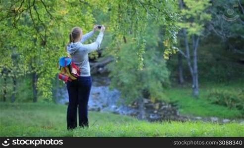 Young woman with backpack taking pictures of scenes in the park or forest using phone camera.