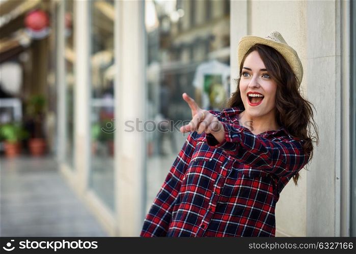Young woman with beautiful blue eyes wearing plaid shirt and sun hat pointing with her finger. Girl in urban background.
