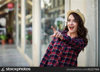 Young woman with beautiful blue eyes wearing plaid shirt and sun hat pointing with her finger. Girl in urban background.