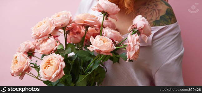 Young woman with beautiful bunch of fresh natural roses coral color in her hands, shoulders with tattoo on a living coral background. Place for text. Postcatd for Woman's or Mother's Day.. Nice fresh bouquet of coral colored roses in girl's hand and tattoo on her shoulder on a living coral background, copy space. Gift for Mother's Day