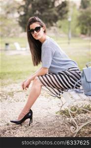 Young woman with beautiful legs in urban background wearing casual clothes. Girl with sunglasses wearing striped skirt and sweater sitting on a bench of an urban park