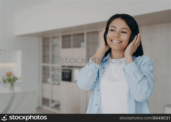 Young woman with beautiful smile is listening to melody in headphones. Carefree spanish girl in earphones is taking pleasure from music. Enjoying playlist in internet. Relaxation and entertainment.. Young woman with beautiful smile is listening to melodies playlist in headphones at home.