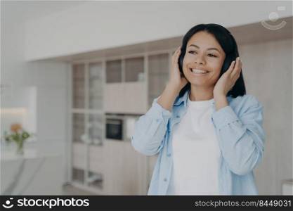 Young woman with beautiful smile is listening to melody in headphones. Carefree spanish girl in earphones is taking pleasure from music. Enjoying playlist in internet. Relaxation and entertainment.. Young woman with beautiful smile is listening to melodies playlist in headphones at home.