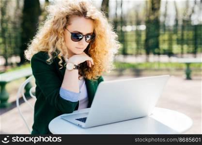 Young woman with blond curly hair wearing sunglasses and elegant green jacket sitting in front of open laptop outdoors reading news online having pensive expression. People, technology concept