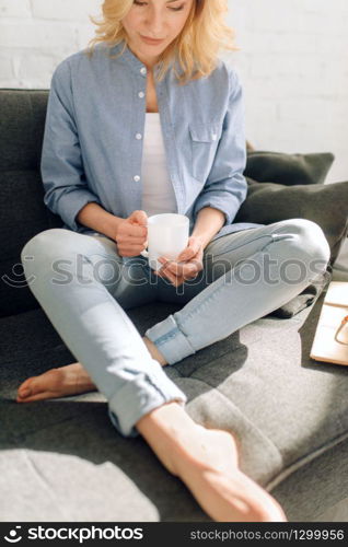 Young woman with book drinks coffee on cozy black couch, living room in white tones on background. Attractive female person with magazine sitting on sofa at home. Woman with book drinks coffee on cozy black couch