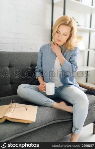 Young woman with book drinks coffee on cozy black couch, living room in white tones on background. Attractive female person with magazine sitting on sofa at home