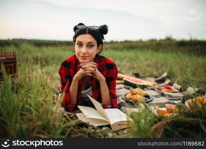 Young woman with book lies on plaid, picnic in summer field. Romantic junket, happy holiday