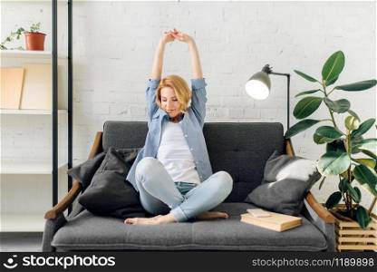 Young woman with book stretch out on cozy black couch, living room in white tones on background. Attractive female person with magazine sitting on sofa at home