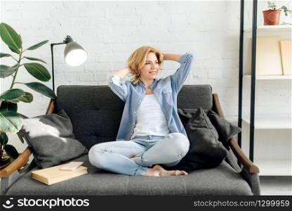 Young woman with book stretch out on cozy black couch, living room in white tones on background. Attractive female person with magazine sitting on sofa at home. Young woman with book stretch out on cozy couch