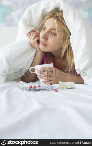 Young woman with coffee mug and medicines suffering from fever while covered in quilt on bed