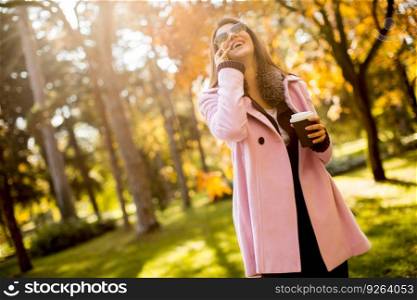 Young woman with coffee to go in hand using a mobile phone and walking in the autumn park