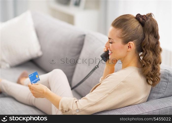 Young woman with credit card talking phone while laying on sofa . rear view