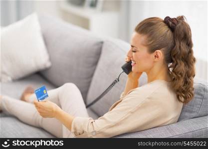 Young woman with credit card talking phone while laying on sofa . rear view