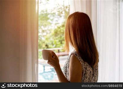 young woman with cup of coffee opening curtains in the morning and looking through the window