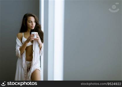 Young woman with cup of coffee or tea in neglige standing by the window in the room