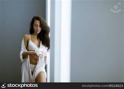 Young woman with cup of coffee or tea in neglige standing by the window in the room