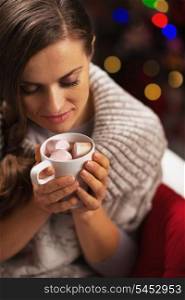 Young woman with cup of hot chocolate with marshmallow in front of christmas lights