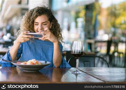 Young woman with curly hair taking photos of her food with a mobile phone while having lunch at a restaurant.