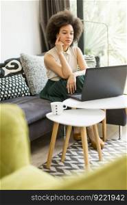 Young woman with curly hair, uses  laptop and sitting on the sofa at home