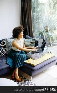 Young woman with curly hair, uses laptop and sitting on the sofa at home