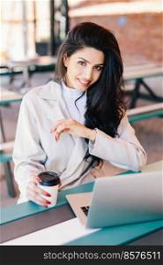 Young woman with dark hair having bright eyes, full lips and healthy skin wearing white coat resting at cafe and browsing internet using laptop computer drinking tasty coffee. Beauty and youth concept