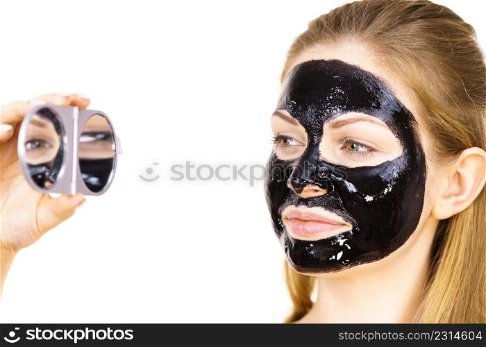 Young woman with detox black peel off mask on face looking in mirror. Spa treatment. Skincare.. Woman with black peel off mask on face